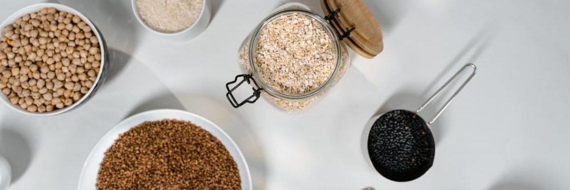 Top view of various grains and seeds in bowls and jars on a white surface.